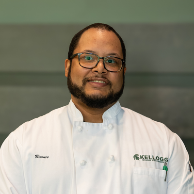 Chef Ronnie Collins faces the camera in a white chef coat reading "Ronnie" on the left of the shirt and "Kellogg" with an MSU Spartan helmet, as well as a green pen in his shirt pocket. Ronnie has a beard and is wearing glasses.