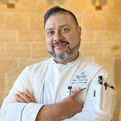 Chef Ben Segueda with arms crossed, wearing a white chef coat, smiling at the camera, standing in front of yellow brick wall.