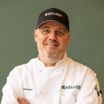 Portrait of Chef Christoph Waldburger wearing a white Kellogg Hotel & Conference Center chef coat and a black Kellogg-branded baseball cap. He is smiling with his arms crossed against a muted green background.